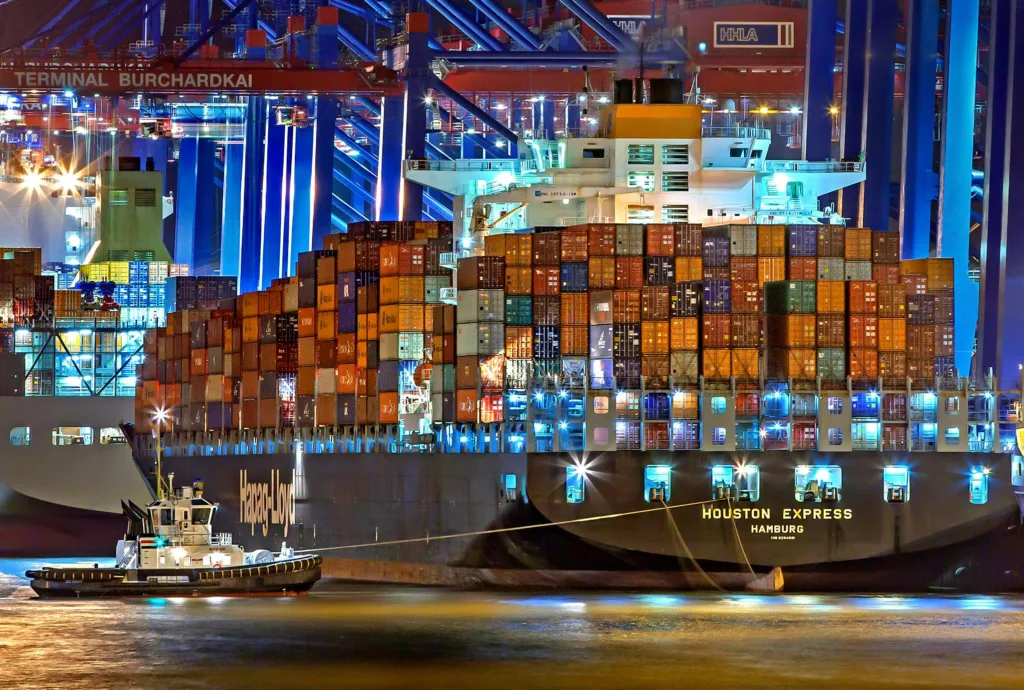 logstics 5 A brightly lit cargo ship at Hamburg harbor with stacked containers and a tugboat.