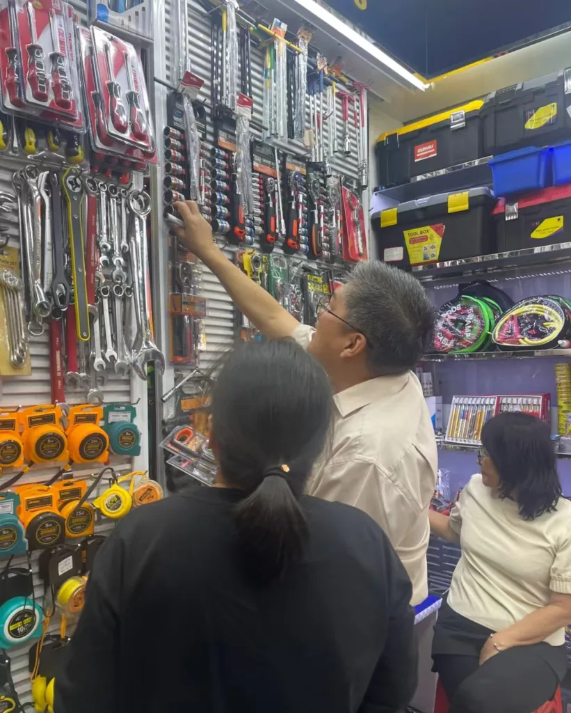 Customers examining hand tools on a wall display inside a hardware store, with wrenches, tape measures, tool kits, and various packaged tools visible.