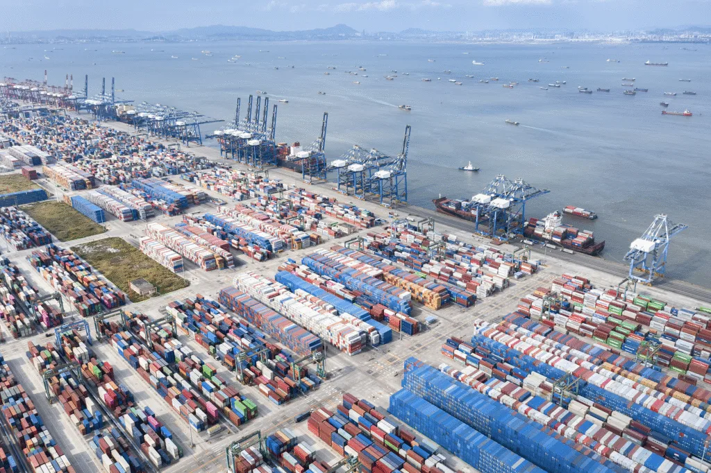 Aerial view of a major shipping port filled with colorful cargo containers, cranes, and docked container ships along the coastline.