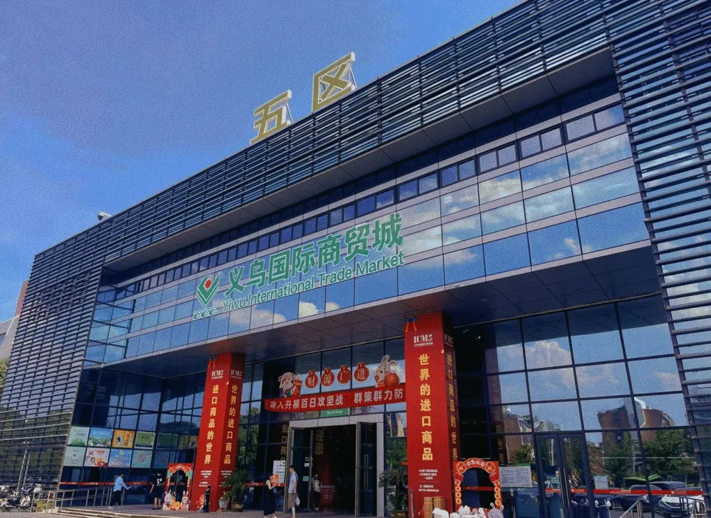Exterior view of Yiwu International Trade Market District 5, showing the modern glass building facade, signage, and entrance decorated with red banners promoting imported goods.