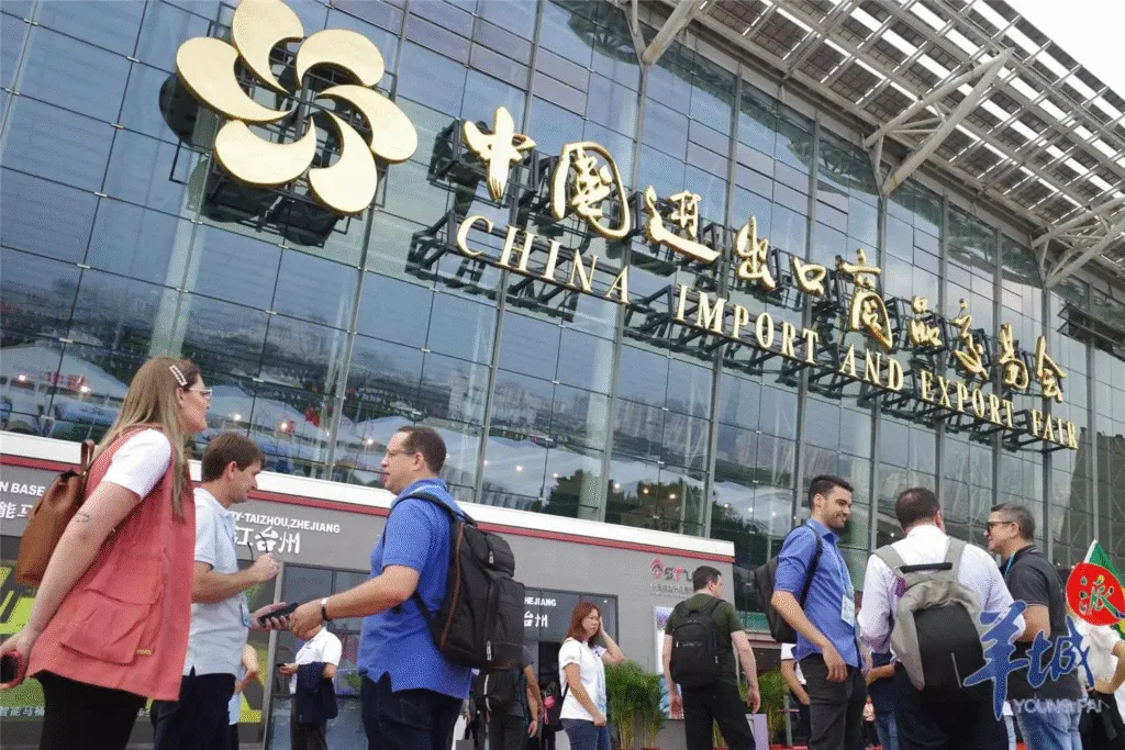 Visitors standing and talking outside the China Import and Export Fair building, with the large gold signage and glass facade of the Canton Fair complex in the background.