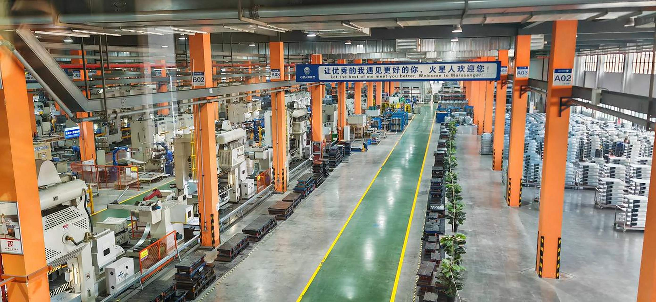 Wide view of an industrial metal stamping factory with heavy machinery, orange support columns, and a central walkway dividing production and storage areas.