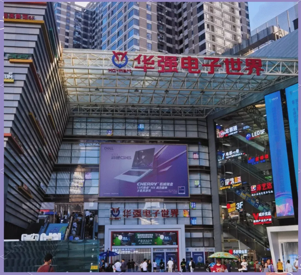 Exterior view of Huaqiangbei Electronics World (HQ-mart) in Shenzhen, showing the large entrance, advertising screens, escalators, and surrounding high-rise buildings with crowds of shoppers.