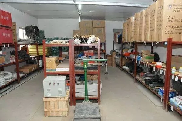 Small warehouse storage area with metal shelving units holding boxes, tools, pipes, and industrial supplies arranged in an organized indoor space.