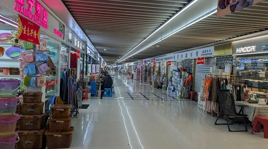 Long indoor wholesale market hallway with small retail shops selling household goods, clothing, and plastic products, illuminated by ceiling lights.