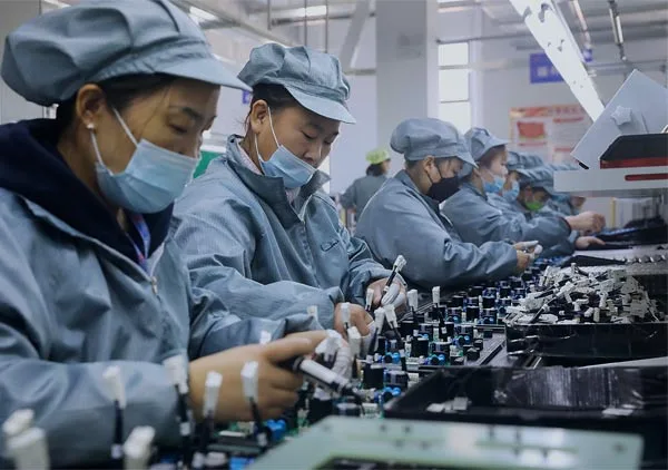 Factory workers wearing protective uniforms and face masks assembling electronic components on a production line in a manufacturing facility.