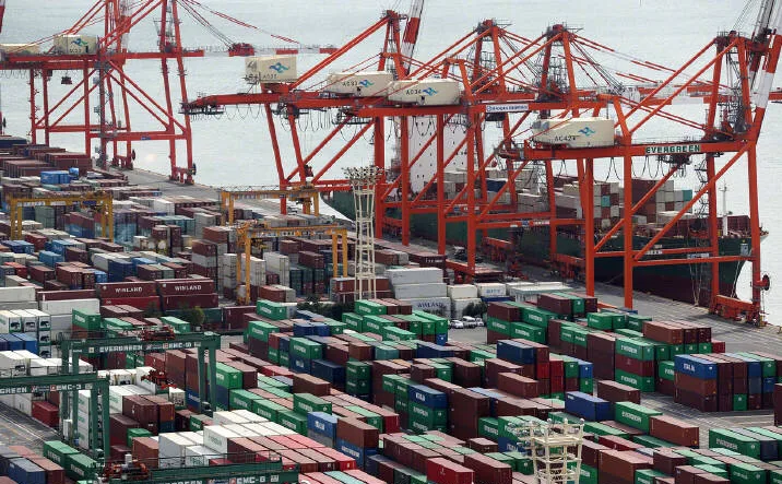 A large shipping port filled with colorful stacked cargo containers and multiple orange loading cranes operating along the waterfront.